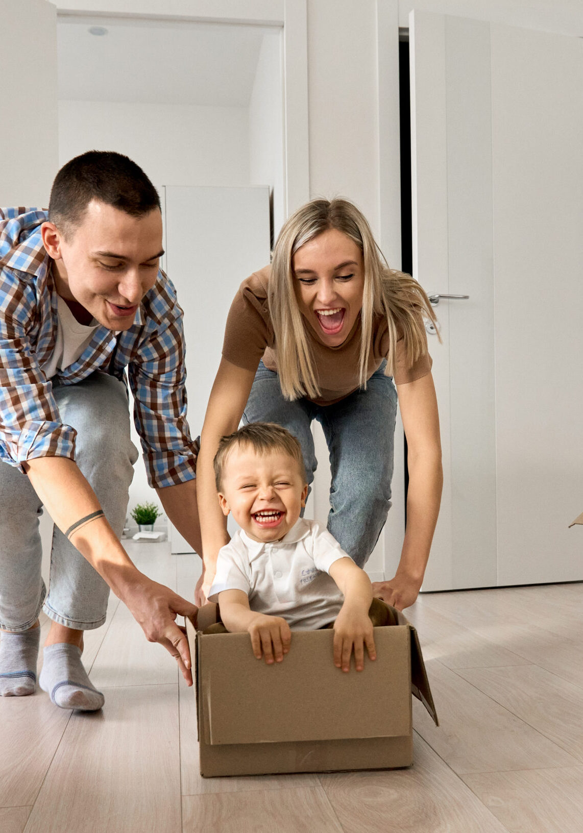 Happy young parents pushing carton box with kid in new mortgage apartment. Overjoyed excited happy householders renters tenants parents pushing carton box with kid riding in new apartment, having fun in their first moving day to real estate property. New home concept.